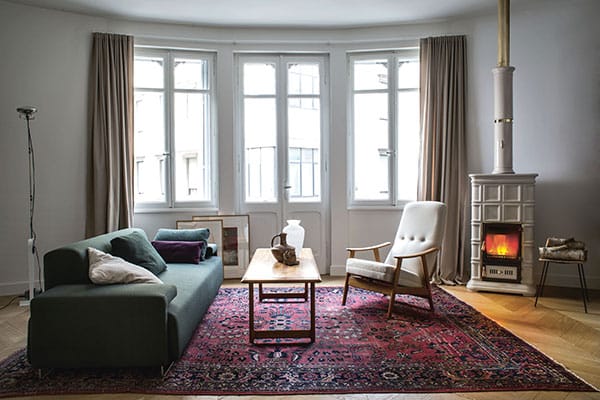 Un salon chaleureux avec un canapé vert, une table basse en bois et un fauteuil couleur crème sur un tapis à motifs rouges. Un poêle à bois Sergio Leoni allumé est placé à droite, à côté d'une table d'appoint. Trois grandes fenêtres avec des rideaux beiges illuminent l'espace.