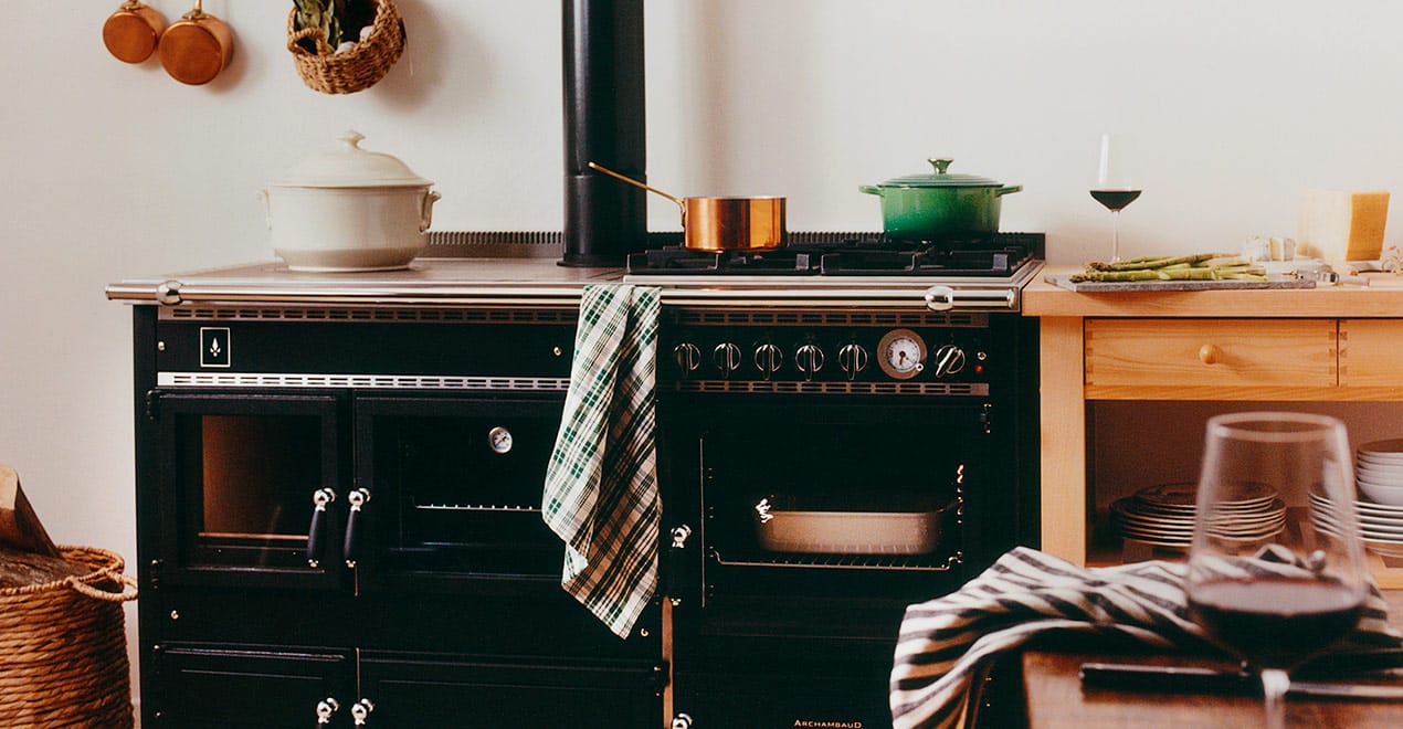 Une scène de cuisine chaleureuse avec une cuisinière Legende noire de style vintage avec une marmite blanche, une petite marmite en cuivre et une marmite verte sur les brûleurs. Une serviette à carreaux est drapée sur la porte du four. Au premier plan, un verre à vin et un torchon sont posés sur le comptoir. Des étagères avec des plats et une bougie sont à l'arrière-plan.