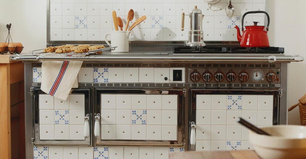 Une cuisine rustique avec un poêle en faïence aux carreaux blancs et bleus, un assortiment d'ustensiles de cuisine dans un support, une bouilloire rouge sur le feu et une plaque de cuisson avec des biscuits sur une serviette. Il y a aussi un bol Origine sur une table en bois au premier plan.