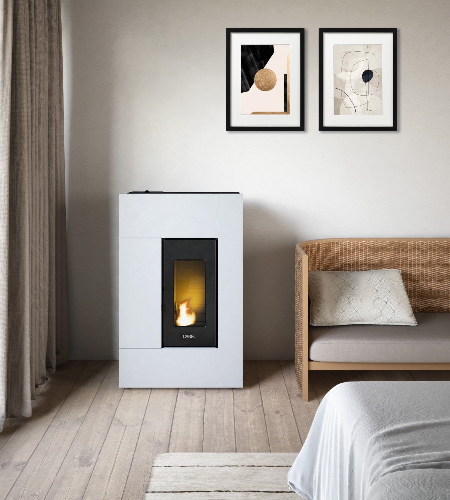 Une pièce moderne est dotée d'un poêle à granulés CADEL Spirit de 5 kW avec une flamme visible, d'une chaise en osier avec un coussin et d'une œuvre d'art abstraite encadrée sur le mur. L'espace présente des murs de couleur claire, un parquet en bois et un rideau partiellement visible sur la gauche.