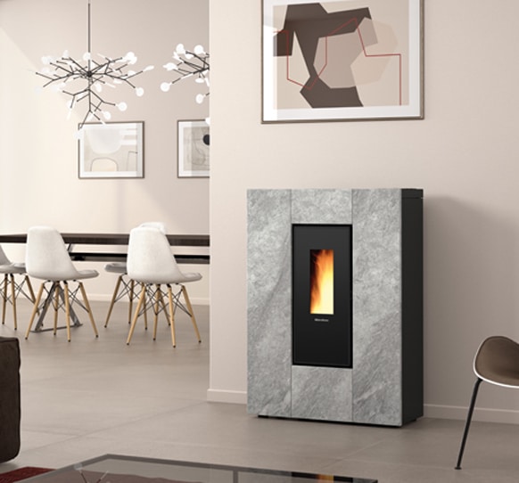 Un espace de vie moderne est doté d'un élégant poêle gris EXTRAFLAME avec un affichage de flamme vertical. La pièce présente un design minimaliste avec des chaises de salle à manger blanches autour d'une table en bois, un luminaire géométrique et des œuvres d'art murales abstraites. Les murs sont beige clair, ajoutant une chaleur subtile au décor inspiré de Marilena.