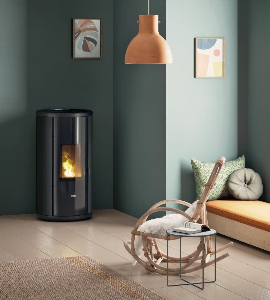 Un salon moderne est doté d'une cheminée cylindrique noire CADEL SOUND 5 PS avec une flamme visible. La pièce a des murs bleu sarcelle ornés d'œuvres d'art abstraites, une lampe en bois, une chaise ronde avec un coussin blanc, une petite table et un banc rembourré le long d'un mur.