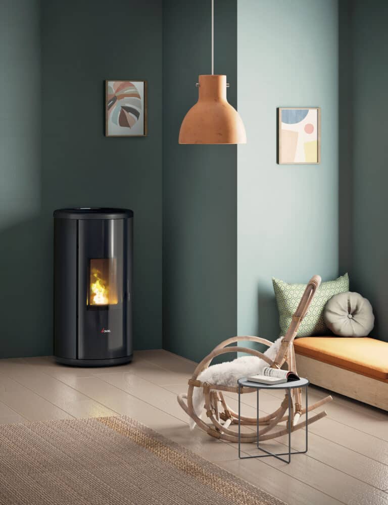 Une pièce moderne aux murs verts abrite un poêle à bois cylindrique noir à flamme visible, peut-être apparenté au poêle à granulés CADEL SOUND 7, une table d'appoint en bois sur laquelle repose un livre, une sculpture beige ressemblant à du bois et un banc rembourré avec des coussins verts. Des œuvres d'art abstraites et une suspension complètent l'espace.