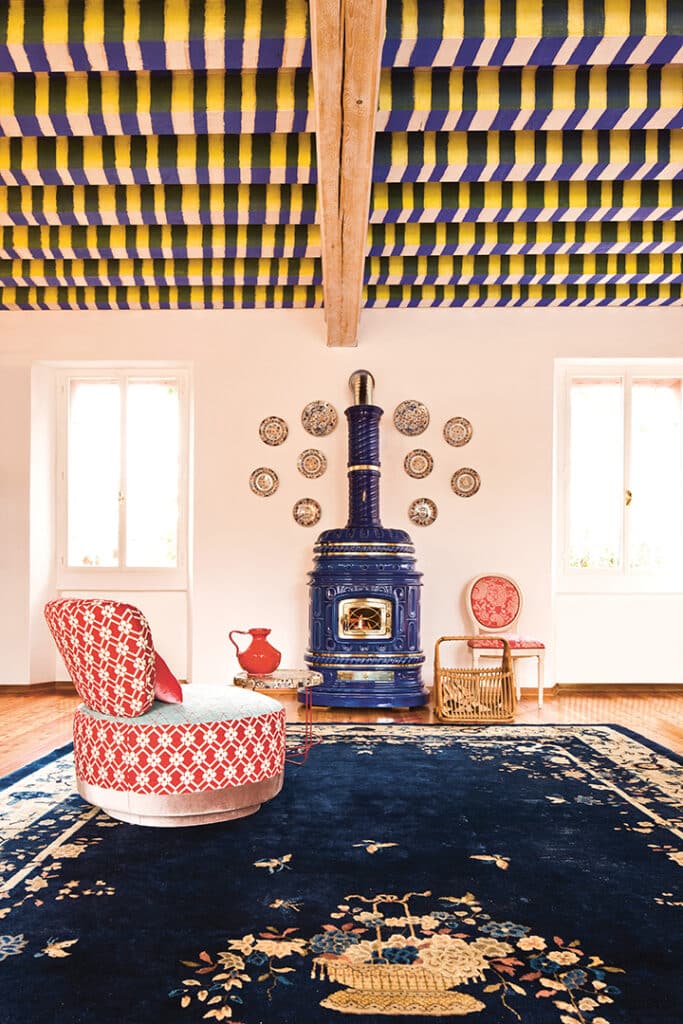 Une pièce au plafond rayé de couleurs, un poêle bleu orné de SERGIO LEONI, un mur orné d'assiettes décoratives, deux chaises (une rouge et une rose) et une petite table avec une cruche rouge. Le grand tapis bleu foncé aux motifs floraux relie l'espace, éclairé par la lumière naturelle à travers deux fenêtres.