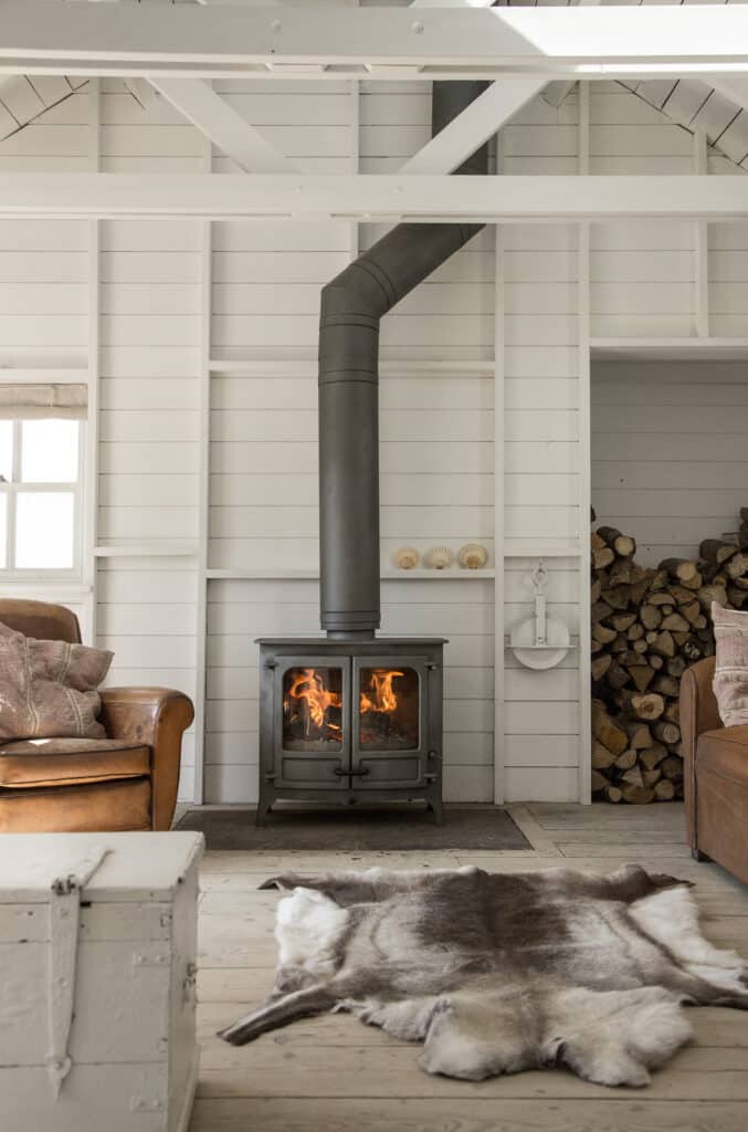 Un salon chaleureux avec un poêle à bois Charnwood Island III et un tuyau qui s'étend jusqu'au plafond. La pièce est dotée de murs en bois blanc, d'un tapis en peau de vache et de fauteuils en cuir. Le bois de chauffage est soigneusement empilé dans une alcôve à côté du poêle. Un coffre blanc fait office de table basse.