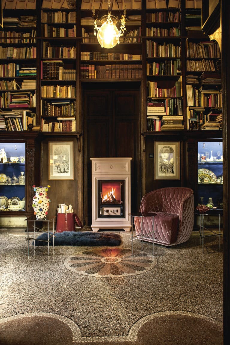 Une pièce chaleureuse avec de hautes étagères remplies de livres, un poêle à bois Sergio Leoni allumé au centre et une chaise rembourrée rose à droite. Le sol est recouvert de carreaux de mosaïque au milieu et des vitrines en verre avec divers objets sont disposées de chaque côté de la cheminée.