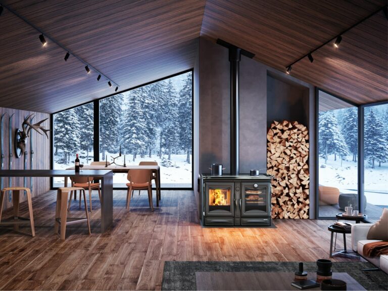 Un intérieur de cabine moderne avec de grandes fenêtres présentant une forêt enneigée, évoquant une sérénité digne de l'ALASKA. Il y a une table à manger avec des chaises, une cuisinière à bois avec un tas de bois de chauffage à côté, et un coin salon confortable avec des fauteuils et une table basse. L'espace présente des murs et des sols en bois.