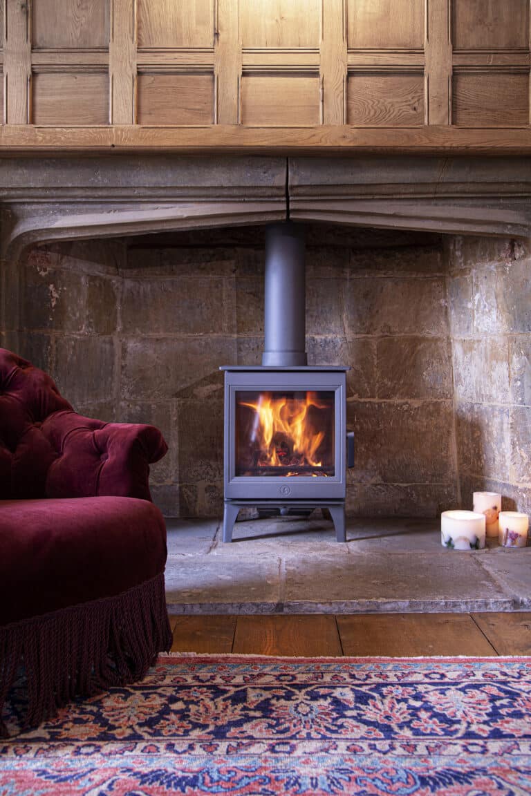 Une pièce chaleureuse est dotée d'un poêle à bois Charnwood Cranmore 5 allumé à l'intérieur d'une cheminée en pierre sous un manteau en bois. À gauche, une chaise rembourrée marron est partiellement visible. Trois bougies allumées sont placées sur le sol près de la cheminée. Un tapis à motifs recouvre le plancher en bois.