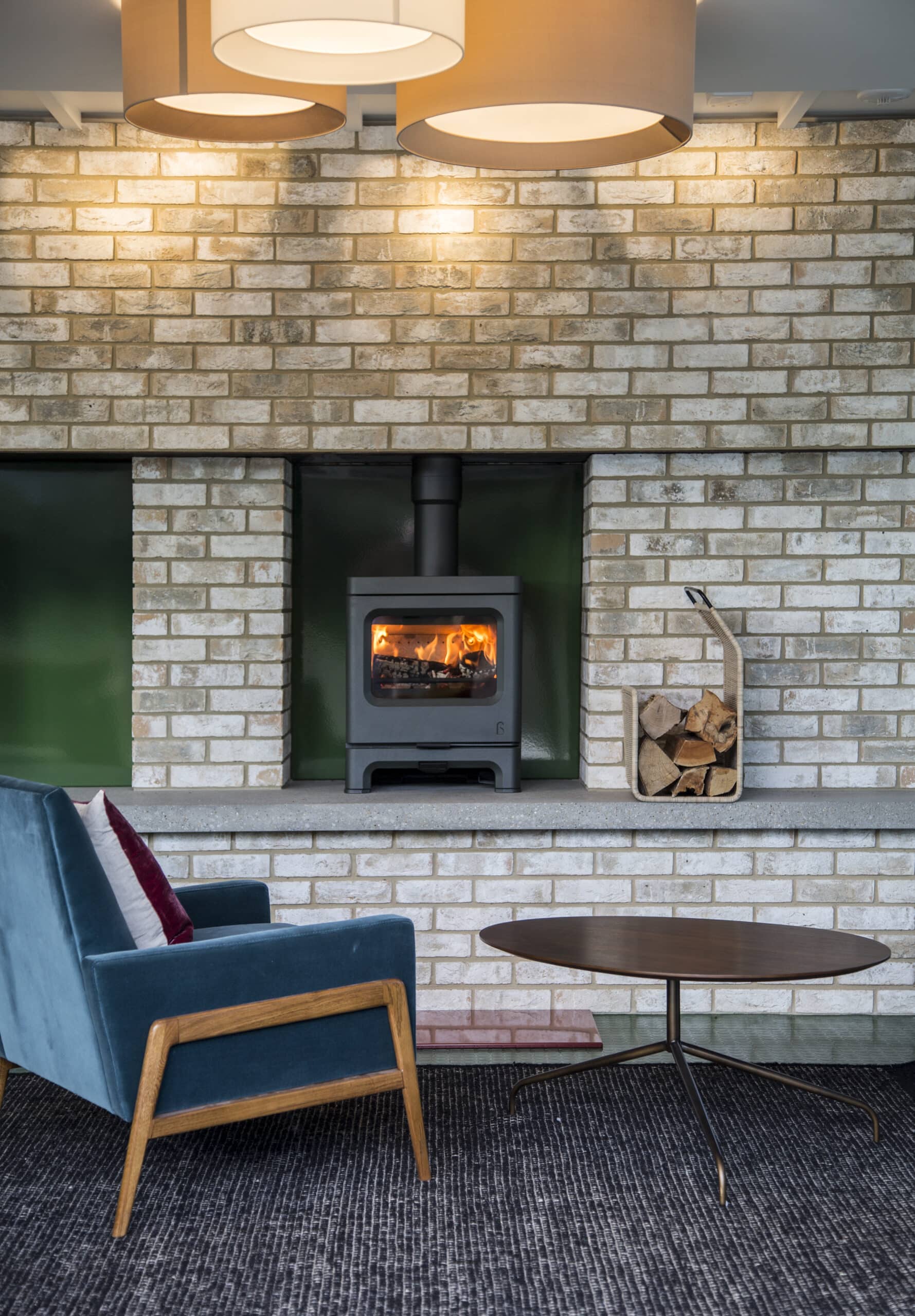 Un espace de vie confortable avec un poêle à bois Charnwood encastré dans un mur en briques. Un fauteuil bleu avec des coussins blancs et bordeaux se trouve à proximité, à côté d'une table basse ronde en bois. Il y a une petite pile de bois de chauffage à côté du poêle Skye 5, et deux grandes suspensions sont suspendues au-dessus.