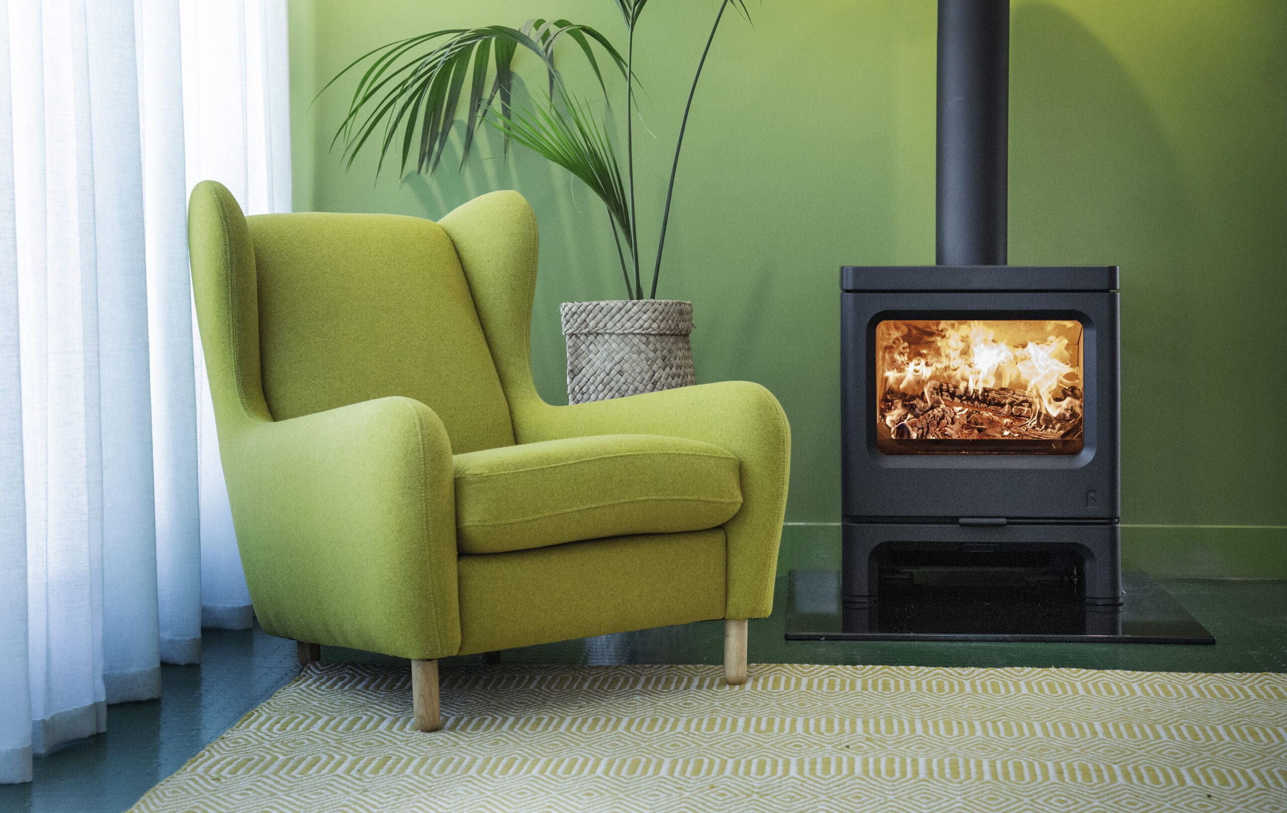 Un fauteuil vert rembourré est posé à côté d'un poêle à bois noir Charnwood Skye 7 avec un feu visible à l'intérieur. Le poêle est placé contre un mur vert clair avec une plante en pot en arrière-plan, et un tapis à motifs de couleur claire est posé sur le sol.