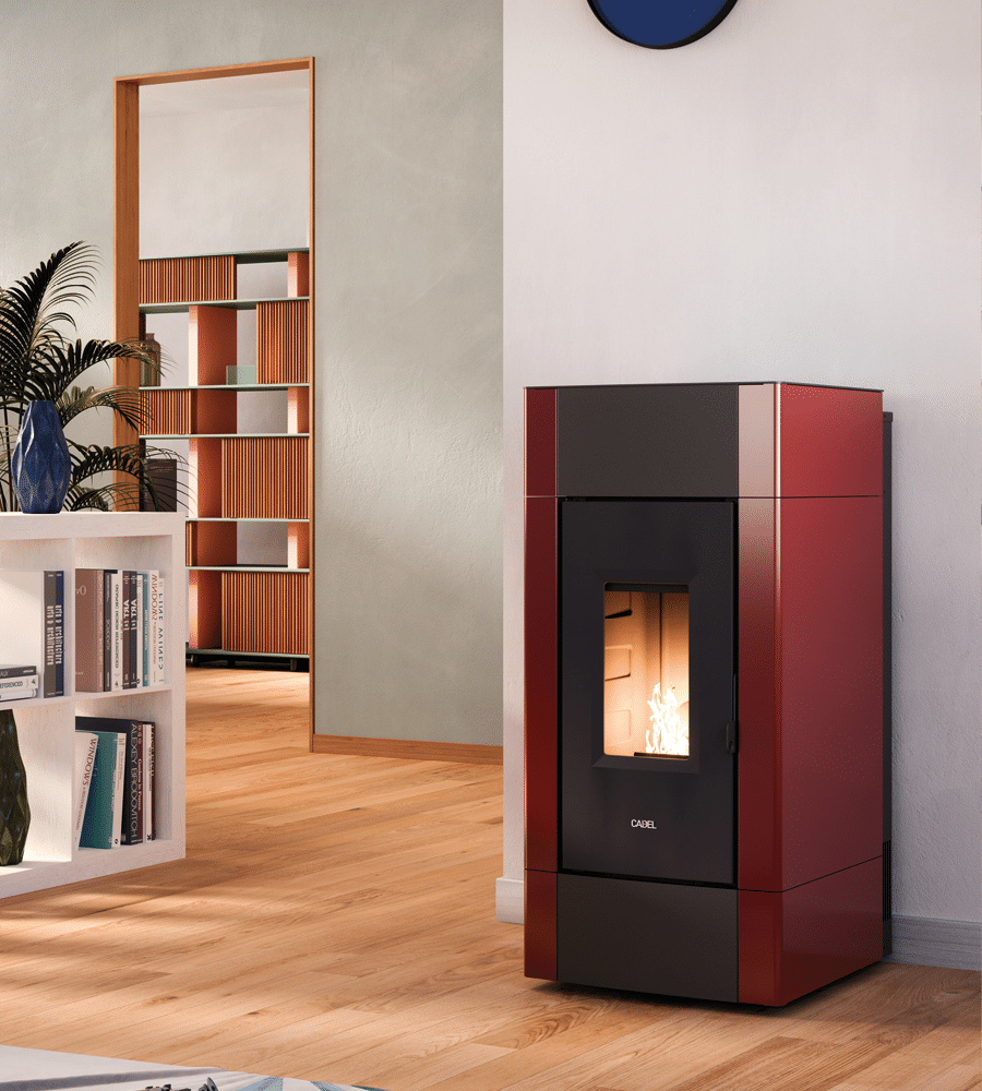 Un poêle à pellets CADEL Cristal Plus 9 rouge et noir trône dans un salon avec une flamme visible à l'intérieur. La pièce est dotée de parquet, d'une plante en pot et d'étagères blanches remplies de livres. Une porte ouverte mène à une autre pièce avec des étagères contre le mur du fond.