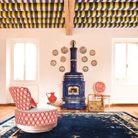 Une pièce au plafond rayé de couleurs, un poêle bleu orné de SERGIO LEONI, un mur orné d'assiettes décoratives, deux chaises (une rouge et une rose) et une petite table avec une cruche rouge. Le grand tapis bleu foncé aux motifs floraux relie l'espace, éclairé par la lumière naturelle à travers deux fenêtres.