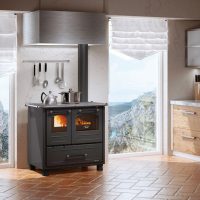 Une cuisine moderne comprend une cuisinière à bois NORDICA FAMILY 4.5 avec deux casseroles sur le dessus, située entre de grandes fenêtres avec vue sur la montagne. Des ustensiles sont suspendus au mur et des armoires en bois avec un comptoir en marbre se trouvent sur la droite. Des stores romains blancs couvrent les fenêtres, ajoutant une touche d'élégance.